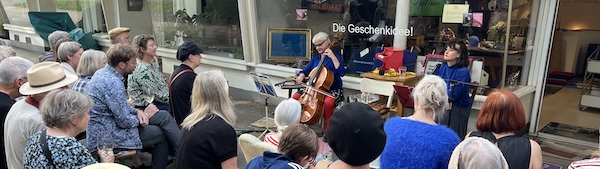 Feierabendlesungen | Dreckstückchen Fußmatten Outdoor street concert by a storefront window, a small string quartet performing for a diverse crowd seated and standing on the sidewalk.