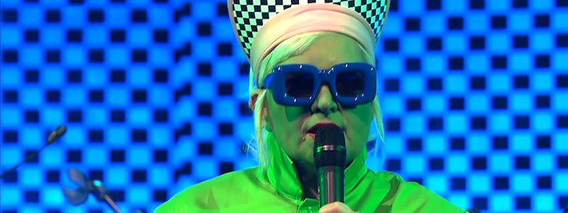 MariolaBrillowska | Dreckstückchen Fußmatten Performer wearing a neon green outfit and blue sunglasses singing into a microphone on a blue checkered stage backdrop.